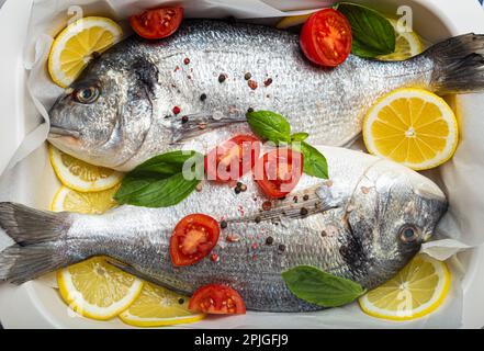 Zwei rohe Fische dorado in einem Auflauf mit Zutaten wie Zitrone, frischem Basilikum und Kirschtomaten aus nächster Nähe auf rustikalem steingrauem Hintergrund Stockfoto