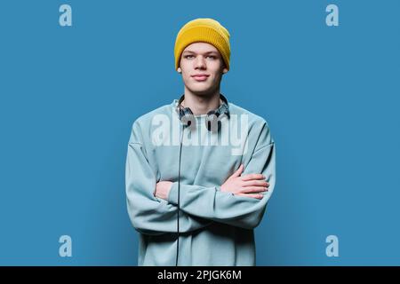 Junger Mann in Sweatshirt mit gelber Kapuze und Kopfhörern auf blauem Hintergrund Stockfoto