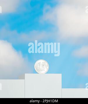 Eine Münze mit einer Stückelung eines russischen Silberrubes steht auf einem Podium vor dem Hintergrund eines blauen Himmels mit Wolken Stockfoto