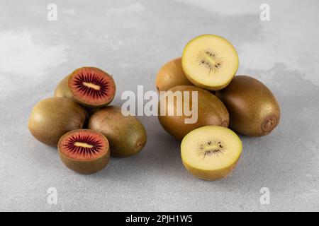 Rote und goldene Kiwi, ganz und halbiert auf einem grauen Tisch. Verschiedene Kiwisorten. Stockfoto