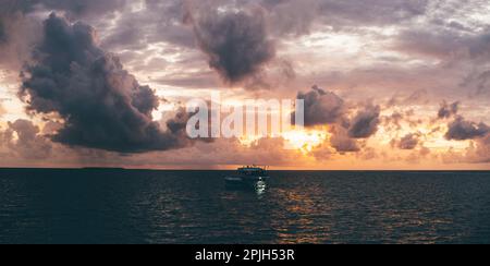 Ein atemberaubender Sonnenuntergang mit unauffälligem Panoramablick auf ein Boot im Meer. Die orangefarbene Sonne ist teilweise hinter violetten und rosafarbenen Wolken verborgen; die Silhouette des Boots Stockfoto