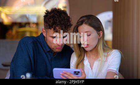 Verärgerter Mann und verärgerte Frau, die im Café sitzen und auf den Bildschirm eines Mobiltelefons schauen. Eine Frau, die ihr Smartphone in der Nähe ihres Freundes hält. Beziehung, Lifestyle-Konzept Stockfoto