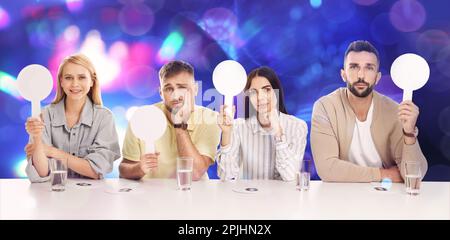 Jury-Gremium mit unterschiedlichen Emotionen, die vor verschwommenem Hintergrund leere Schilder am Tisch halten. Bokeh-Effekt Stockfoto