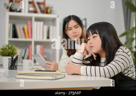 Ein faules und gelangweiltes, junges asiatisches Mädchen sitzt an einem Tisch mit der Hand am Kinn, zu müde, um zu lernen oder Hausaufgaben zu machen. Stockfoto