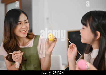 Lachende und fröhliche asiatische mutter, die ihrer kleinen Tochter dabei hilft, ein Osterei zu malen und zu Hause Spaß miteinander zu verbringen. Stockfoto