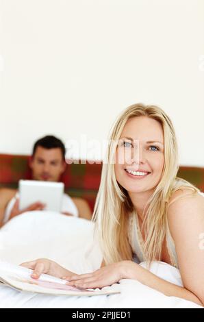 Wir machen eine Pause von unserem hektischen Lebensstil. Porträt einer jungen Frau, die im Bett liest, während ihr Mann im Hintergrund liegt. Stockfoto