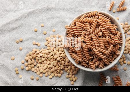 Grüne Fusilli-Nudeln mit Linsen auf grauem Textilhintergrund. Eine Schüssel mit roher Pasta und grünen Linsen. Glutenfreie Pasta. Stockfoto