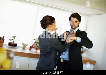 Die Zeit im Auge behalten. Eine attraktive Frau repariert ihren Mann am Kragen, während er auf seine Uhr schaut. Stockfoto