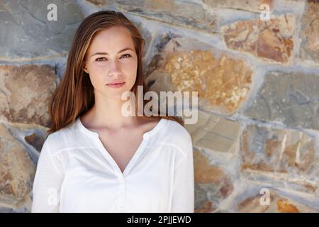 Ich genieße die Ruhe, allein zu sein. Eine junge Frau steht draußen und lehnt sich gegen eine Steinmauer. Stockfoto