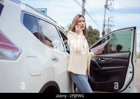 Unzufriedene Geschäftsfrau, die neben einem Auto steht und ein Dokument hält und ein Mobiltelefon benutzt. Stockfoto