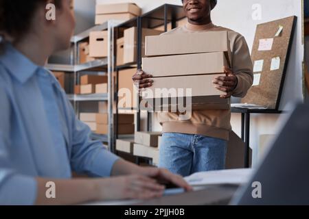 Konzentrieren Sie sich auf den jungen schwarzen Mann, der einen Stapel verpackter Kartons mit sich führt, während er auf seinen Kollegen zugeht und dabei neue Online-Bestellungen entgegennimmt Stockfoto