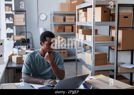 Seriöser junger Mann, der auf einen Laptop-Bildschirm schaut, während er am Arbeitsplatz im Lager oder Postamt sitzt und Daten zu Online-Bestellungen prüft Stockfoto