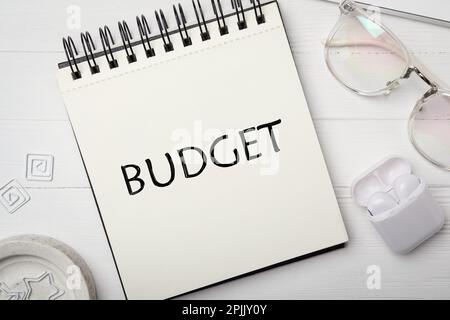 Notizbuch mit Wort Budget, Brille und Ohrhörer auf weißem Holztisch, flach liegend Stockfoto