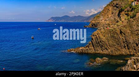 Tagsüber Panoramablick auf das Mittelmeer mit Booten und Klippen über dem Wasser von der Altstadt von Riomaggiore in Cinque Terre, Italien Stockfoto