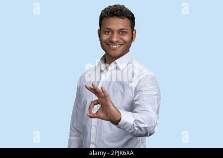 Das Porträt eines jungen indischen Mannes zeigt das OK-Zeichen. Isoliert auf pastellblauem Hintergrund. Stockfoto