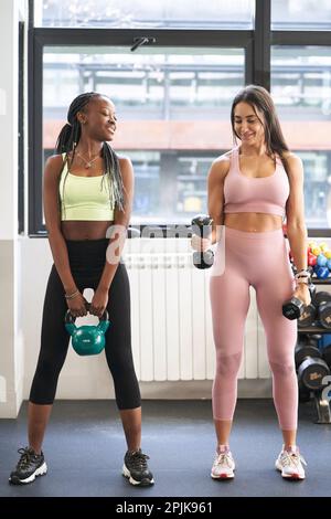 Zwei multiethnische weibliche Freunde trainieren mit Gewichten und haben eine tolle Zeit im Fitnessstudio. Stockfoto