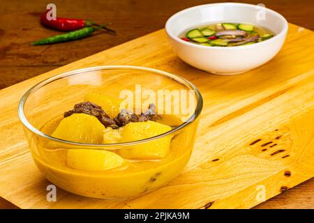 Nahaufnahme von köstlichen hausgemachten Kartoffeln, Rindfleisch, Zwiebeln, Chili und Kokosmilch-Curry in einer transparenten Glasschüssel auf Holzbrett mit einer Tasse Cu Stockfoto