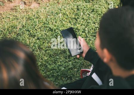 Blick von der hinteren Gruppe lachender Latinos, die auf dem Boden in einem Park mit einem Smartphone sitzen Stockfoto