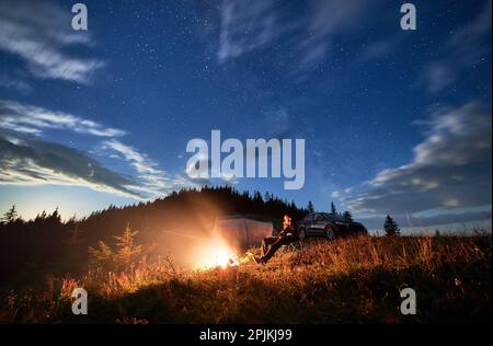 Camping in den Bergen unter bewölktem Nachthimmel. Ein junger Mann ruht sich am Lagerfeuer aus, in der Nähe eines Touristenzelts und eines Geländewagens auf einem Hügel. Ein reisender Mann, der nachts die Sterne beobachtet. Stockfoto