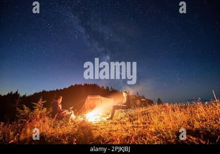 Nachtcampen in den Bergen unter Sternenhimmel voller Sterne. Reisendes Paar, das sich am Lagerfeuer, im Touristenzelt und im Geländewagen am Abend auf dem Rasen der Berge erholt und die Sterne zusammen betrachtet. Stockfoto