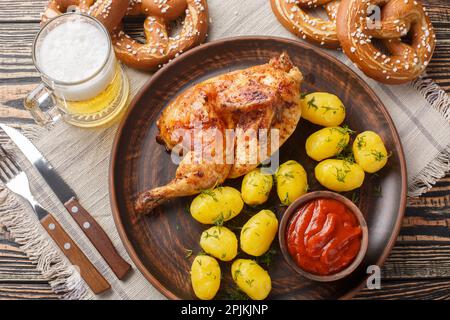 Traditionelles bayerisches Gericht gebratenes halbes Hähnchen mit gekochten Kartoffeln und Tomatensauce serviert mit Bier und Brezel Nahaufnahme auf dem Holztisch. Horizontal Stockfoto