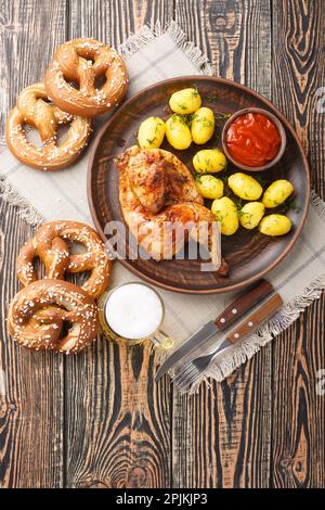 Bayerisches Brathähnchen Hendl mit gekochten Kartoffeln, serviert mit Bier und Brezeln auf dem Holztisch. Vertikale Draufsicht von oben Stockfoto