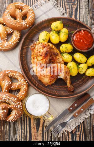 Deutsches Festtagsessen, halb gebratenes Hähnchen mit gekochten Kartoffeln, Soße, Bier und Brezeln, dicht auf einem Holztisch. Vertikale Draufsicht von oben Stockfoto