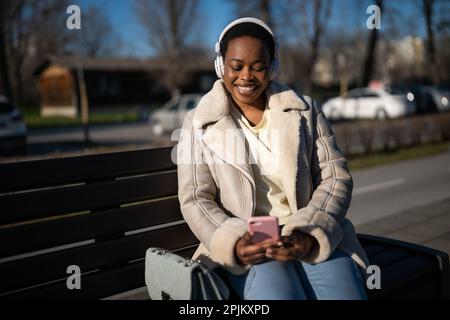 Außenporträt einer glücklichen afroamerikanischen Frau am sonnigen Tag. Sie sitzt auf einer Bank auf der Straße und benutzt ihr Smartphone. Stockfoto