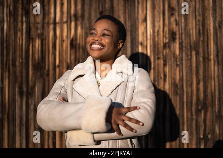 Außenporträt einer glücklichen afroamerikanischen Frau. Sie steht vor einer hölzernen Mauer und lächelt. Stockfoto