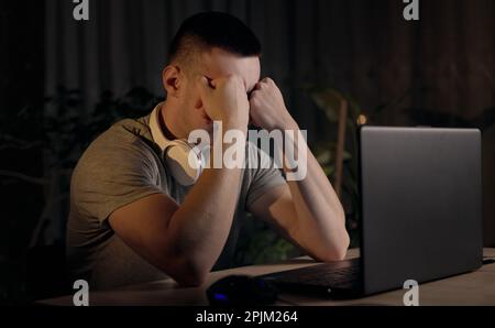 Schläfriger, müder junger Student, der zu Hause spät am Laptop lernt und seine trockenen, gereizten Augen massiert. Überarbeiteter Freiberufler arbeitet an Stockfoto