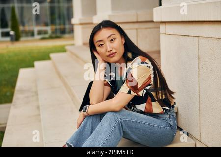 Er sitzt auf der Treppe des Gebäudes. Eine junge Asiatin ist tagsüber draußen Stockfoto