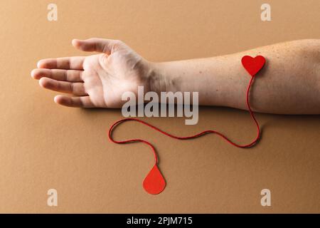 Arm einer weissen Frau mit Herz und roter Schnur mit Bluttropfen auf braunem Hintergrund Stockfoto