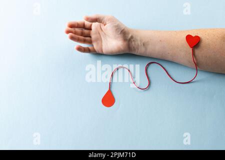 Arm einer weissen Frau mit Herz und roter Schnur mit Bluttropfen auf blauem Hintergrund Stockfoto