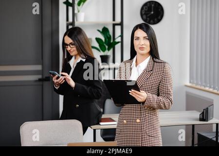 Zwei attraktive junge Frauen arbeiten im Büro. Eine Frau telefoniert und ihr Kollege macht sich Notizen in einem Notizbuch. Stockfoto