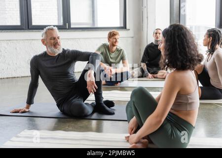 Positiver reifer Mann, der mit einer Frau aus dem Nahen Osten im Yoga-Kurs spricht, Stock Image Stockfoto