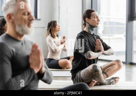 Tätowierter Mann, der anjali Mudra praktiziert, in der Nähe multiethnischer Leute in der Yoga-Gruppe, Börsenbild Stockfoto