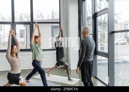 Trainer mittleren Alters, der neben einer multiethnischen Gruppe steht und im Yoga-Studio den Halbmondschritt praktiziert, Stockbild Stockfoto