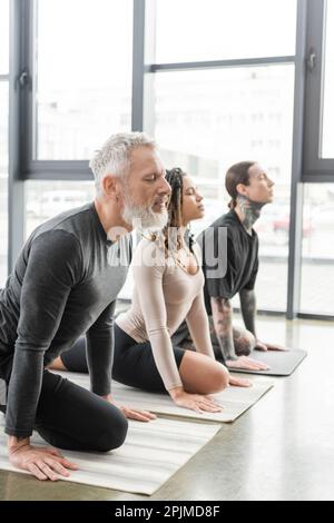 Ein Mann mittleren Alters, der halb Tauben asana praktiziert, in der Nähe multiethnischer Leute im Yoga-Kurs, Stock Image Stockfoto
