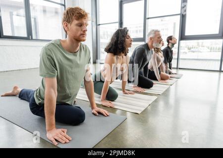 Rothaariger Mann praktiziert Half Pigeon Asana in der Nähe multiethnischer Leute im Yoga-Kurs, Stock Image Stockfoto