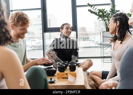 Fröhlicher Mann mit Tattoos, der während der Teezeremonie in der Nähe von zwischenrassischen Freunden im Yoga-Studio saß, Stockbild Stockfoto