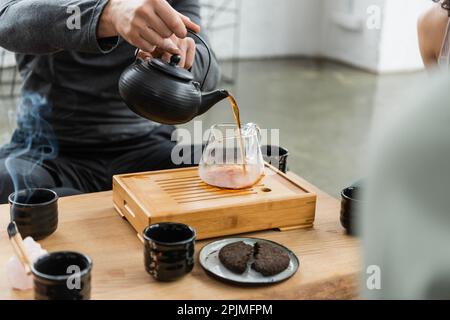 Zugeschnittene Ansicht von Mann, der frisch gebrühten Puer-Tee in eine Glaskanne gießt, Stockbild Stockfoto