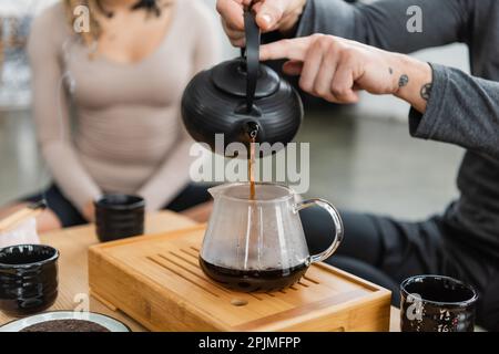 Zugeschnittene Ansicht eines tätowierten Mannes, der frisch gebrühten Puer-Tee in eine Glaskanne in der Nähe einer Frau gießt, auf unscharfem Hintergrund, Stockbild Stockfoto