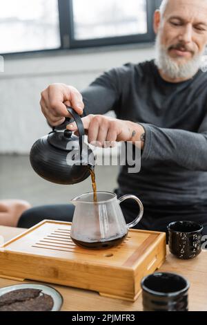 Tätowierter Mann mittleren Alters, der frisch gebrühten Püretee in eine Glaskanne gießt, Stockbild Stockfoto