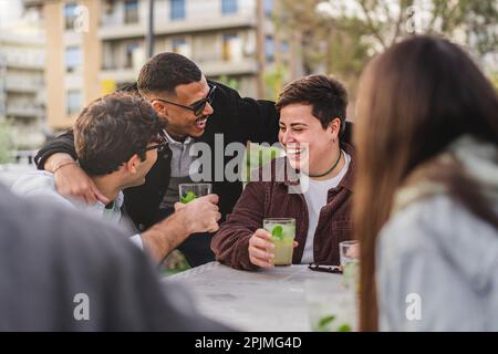 Eine vielfältige Gruppe von Freunden, darunter eine nicht binäre Person in Übergröße und ein weißer Mann, genießen Mojitos in einem Pub im Freien, während sie gerne einen gemischten begrüßen Stockfoto