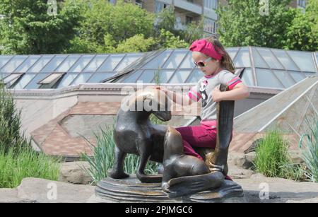 Moskau, Russland, Mai 2012: Ein süßes, modisches Kleinkind sitzt auf einer Messingskulptur einer Navy Seal mit einem Jungen und streichelt es. Stockfoto