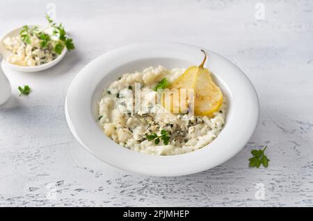 Köstliches hausgemachtes Birnen- und Blaukäse-Risotto mit Petersilie in weißer Schüssel auf graublau strukturiertem Hintergrund Stockfoto