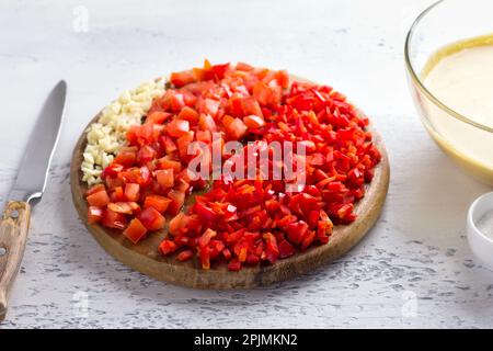 Holzbrett mit gehacktem Gemüse: Rote Paprika, Tomaten, Knoblauch. Kochen von veganem Kichererbsenomelett auf hellgrauem, strukturiertem Hintergrund Stockfoto