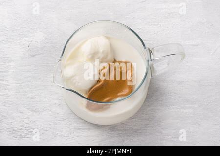 Glaskanne mit Milch, Vanilleeis und Schlagsaum auf hellgrauem Hintergrund. Zubereitung eines trendigen Dalgona-Kaffeegetränks Stockfoto