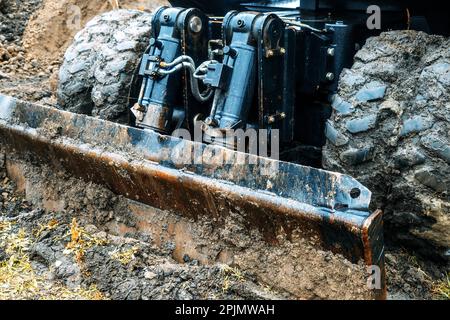 Radtraktor mit Schaufel räumt an bewölkten Regentagen den Boden frei. Schmutzige Bulldozer-Schaufel Nahaufnahme... Stockfoto