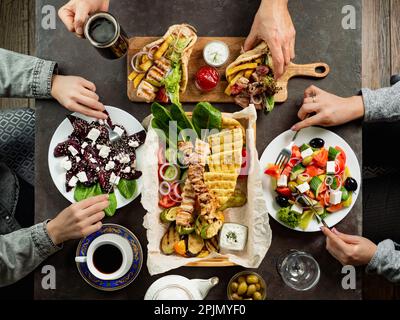 Menschen, die griechisches Essen essen. Griechische Küche auf dunklem Tisch, Blick von oben. Hände mit Gyros, griechischer Salat, Souvlaki - Auswahl an griechischer Küche Stockfoto